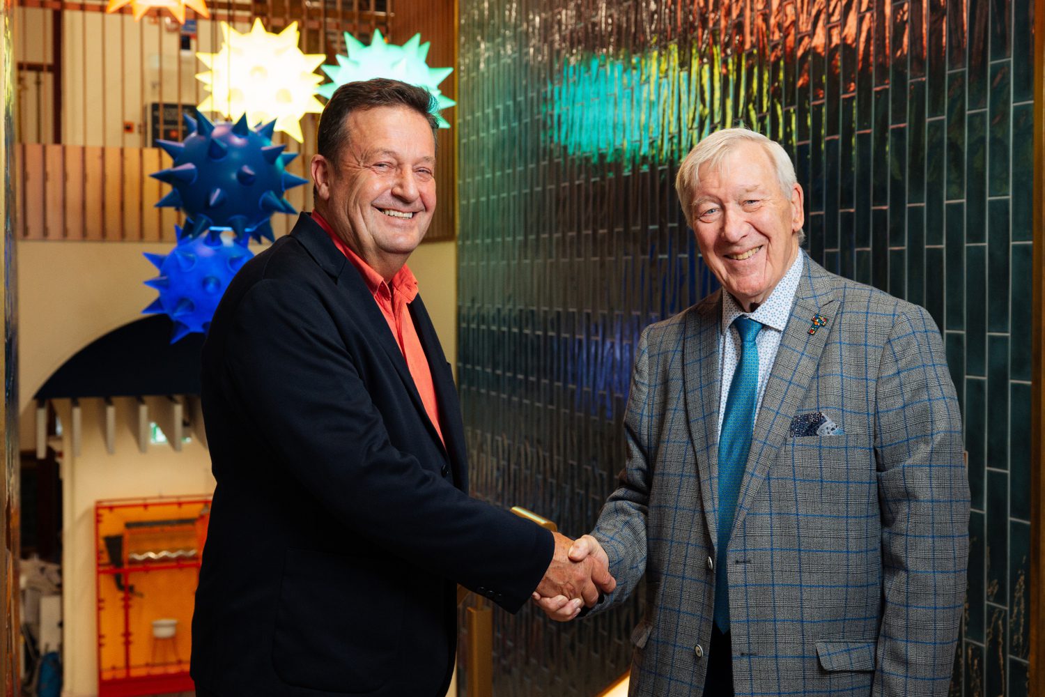 Two men in suits shake hands in a corridor lined with green tiles